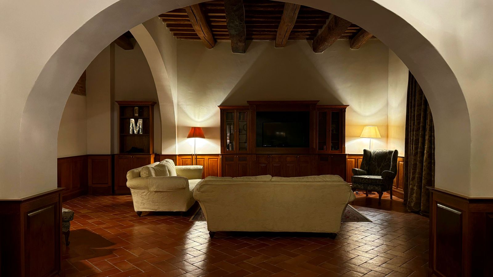 Salotto di una villa toscana con pavimento in cotto antico, soffitto a travi e lampade da tavolo accese